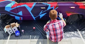 Man with red hair and a red, white, and blue plaid shirt, sits on the curb spray-painting the side of a car with purple, blue, black, and red paint.