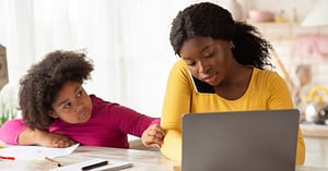 African-American mom wearing a yellow top, talks on her phone while working from home. She is typing on her computer. Her daughter, wearing a pink top with paper and pens around her tries to get her mom's attention.