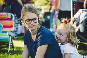 Toddler girl with blonde hair and wearing a white shirt hides behind her mother's back at a park. A folding chair in the background. Mom is wearing a blue t-shirt and glasses and has a serious expression on her face.