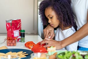 Young African-American girl with curly natural hair learning to cut red bell peppers. Her mother's hands are visible helping her to hold the knife. Photo by Kampus Productions for Pexels.