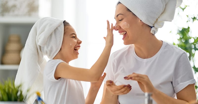 Mother and daughter, with white towels on their hair, apply face mask to one another while smiling.