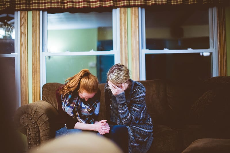 Blond man sits on a couch with a red-haired woman. Both have their heads down in grief. Photo by Ben White for Unsplash.